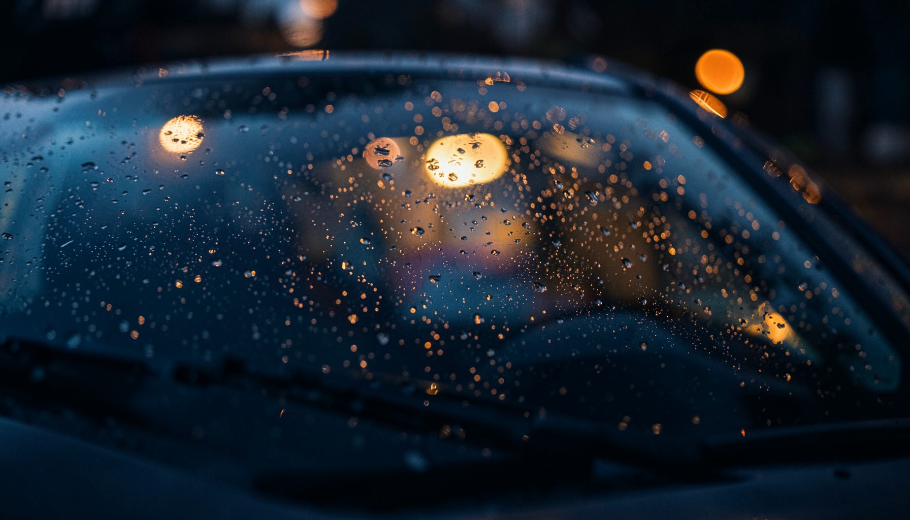 A rain-speckled windshield on a dark vehicle, soft bokeh lights glowing in the background, deep navy and warm amber tones