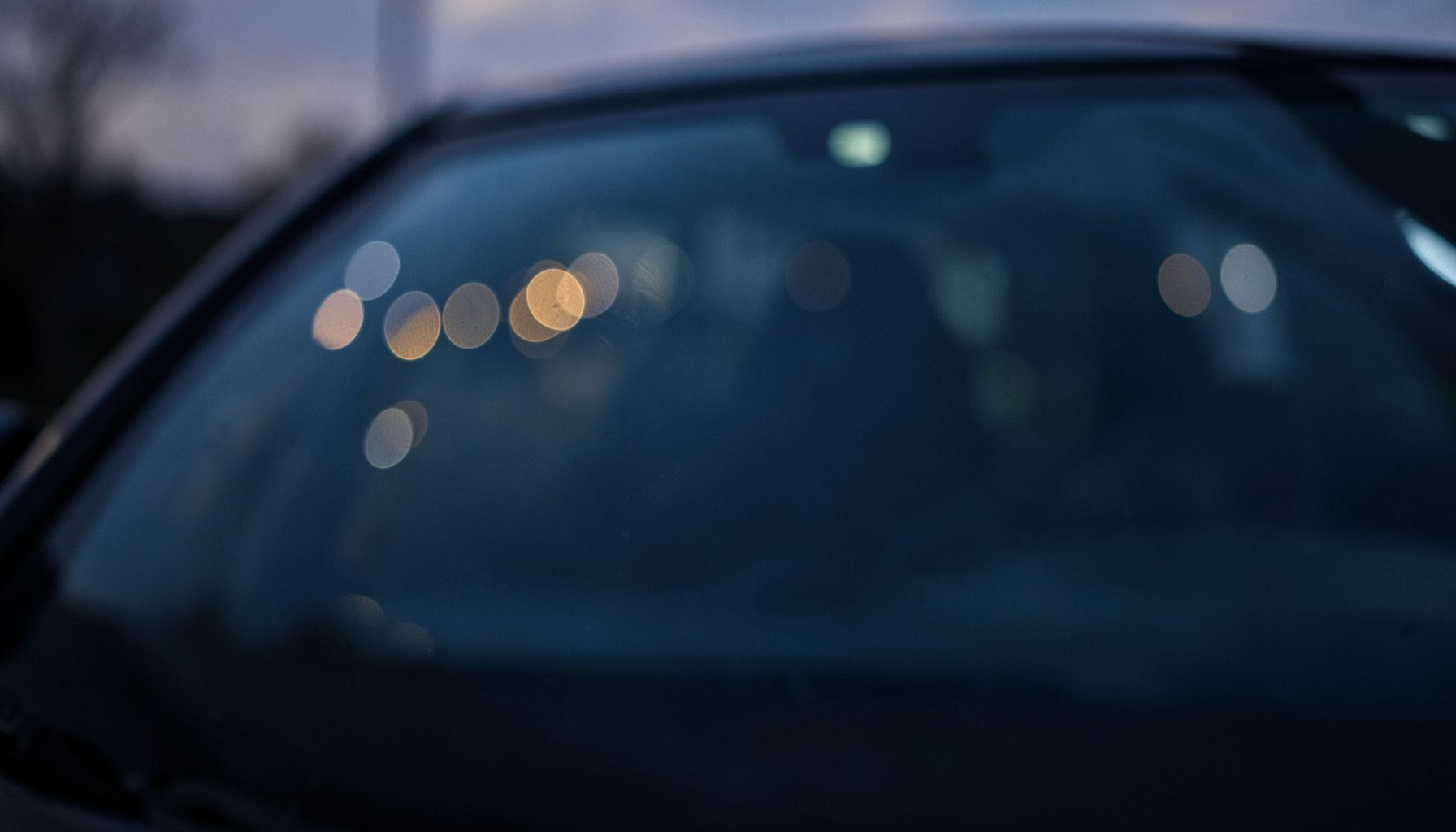 Dark atmospheric close-up of a car windshield with soft bokeh lights reflecting off the glass at dusk, deep navy and charcoal tones