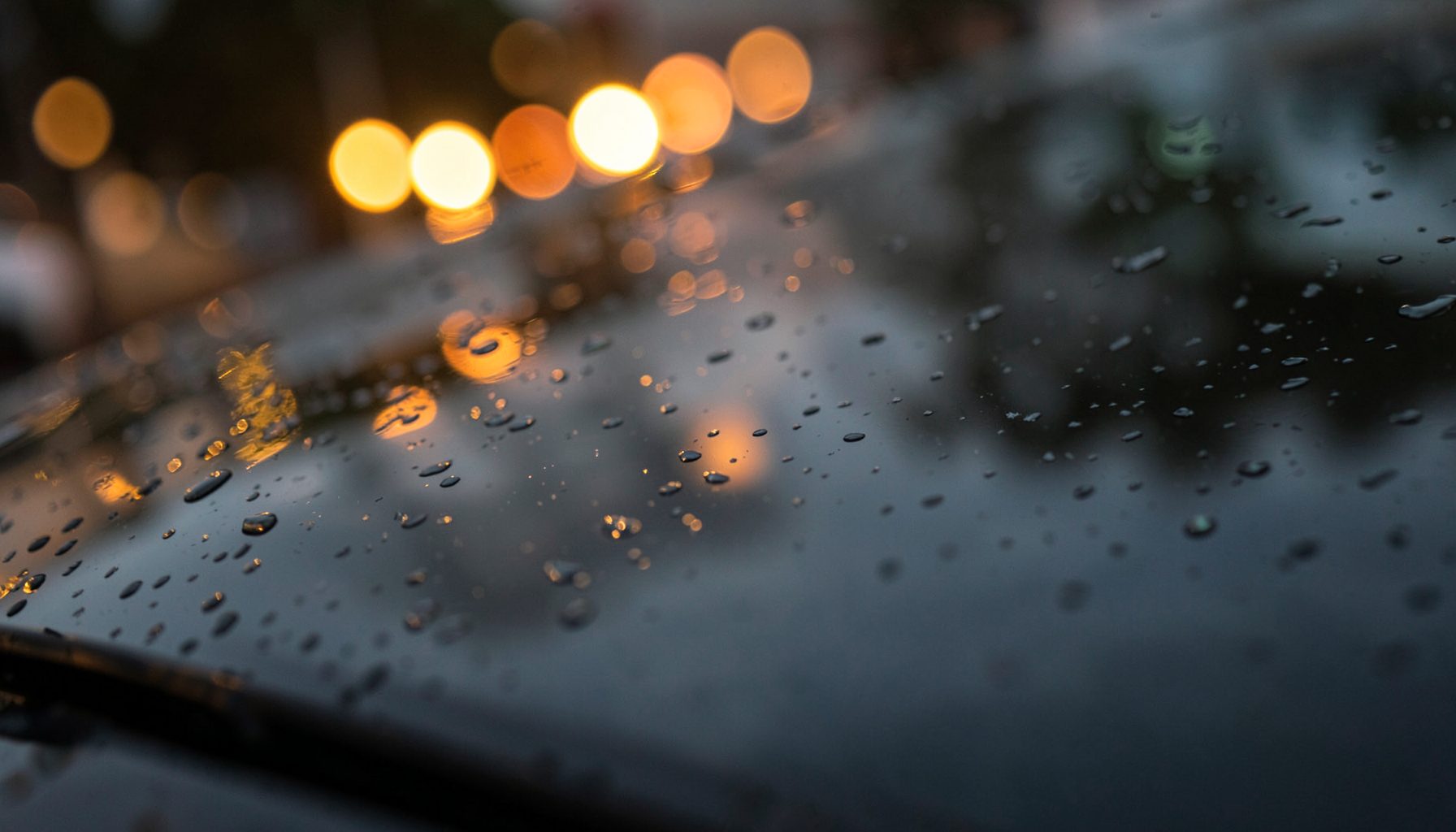 Glossy dark windshield surface with soft rain droplets and warm amber bokeh lights in the background