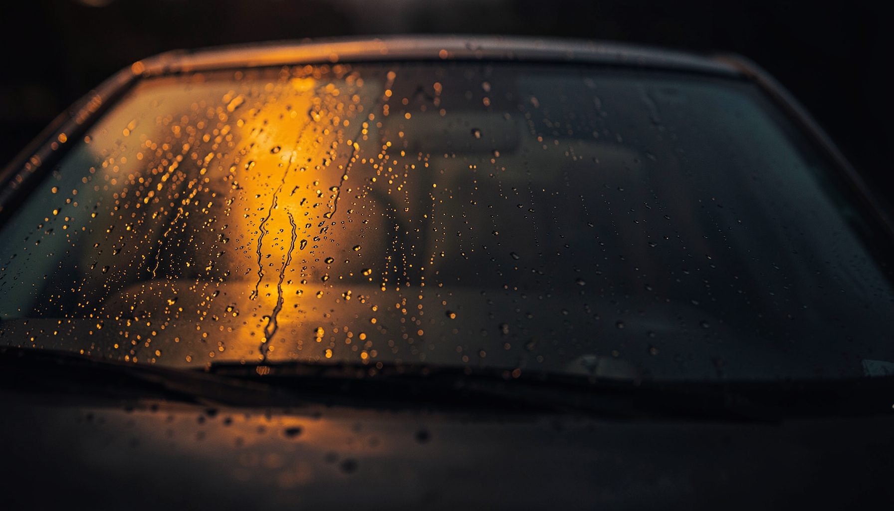 Wet car windshield with water droplets catching warm amber light, dark moody background