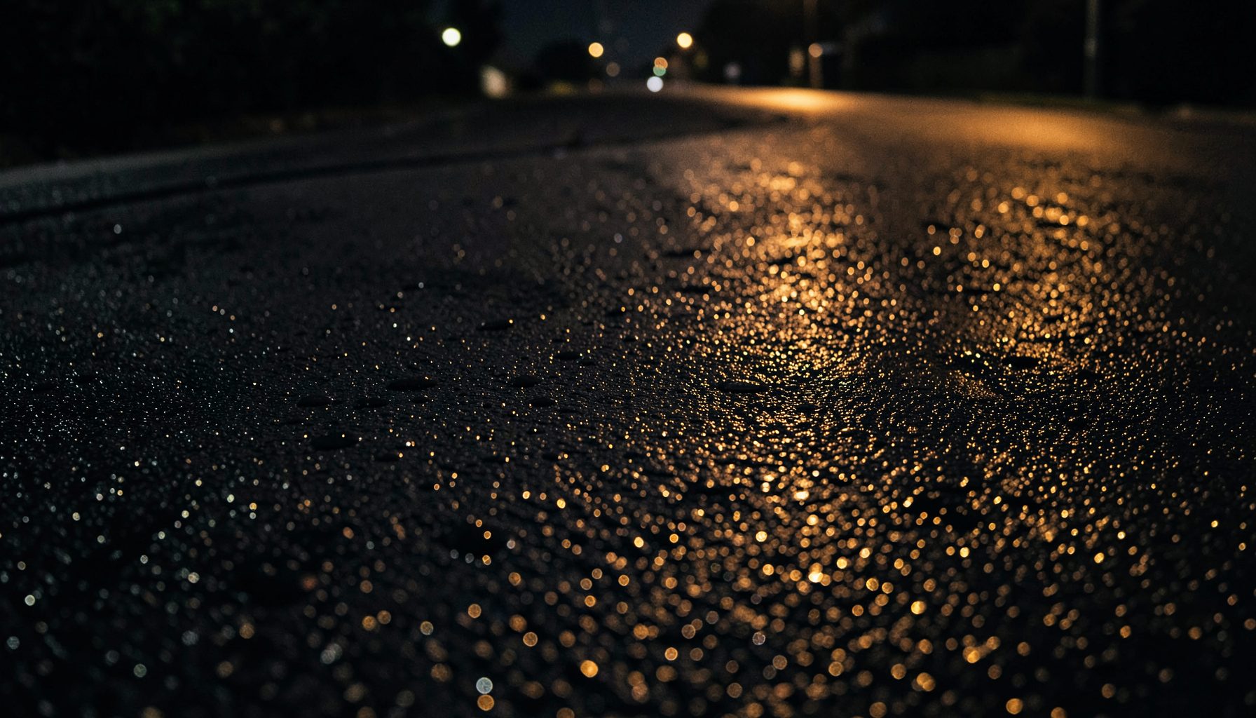 Dark wet asphalt with scattered water droplets reflecting soft amber streetlight glow, moody nighttime atmosphere